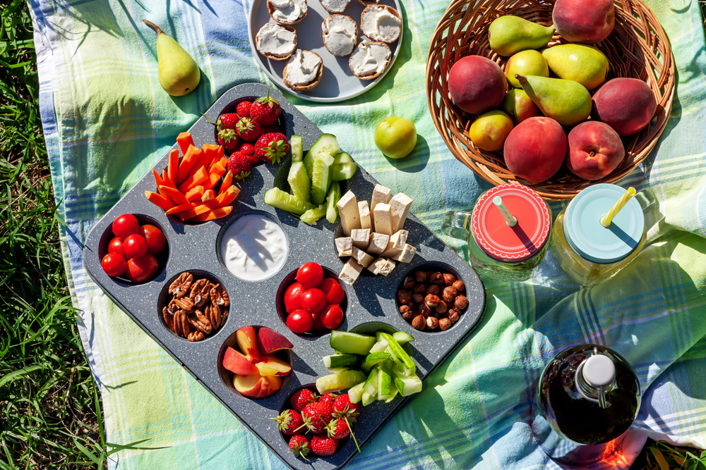Summertime healthy picnic with fresh vegetables, nuts and fruits served in the muffin pan, refreshing drink in mason jar and some snack on blue cloth in the garden